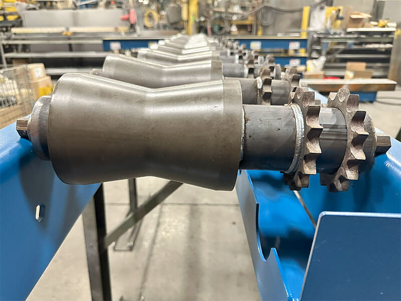 Bowtie-shaped sprocketed rollers mounted in a blue frame for centering cylindrical parts on a CDLR conveyor.